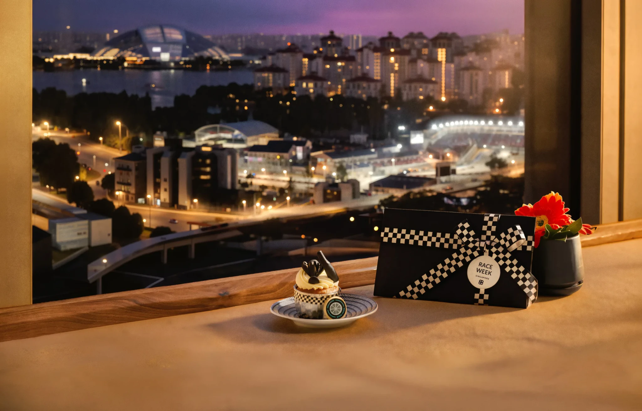 A cupcake with chequered decoration beside a black Race Week gift box and a red flower, with the Marina Bay Street Circuit lit up through the window