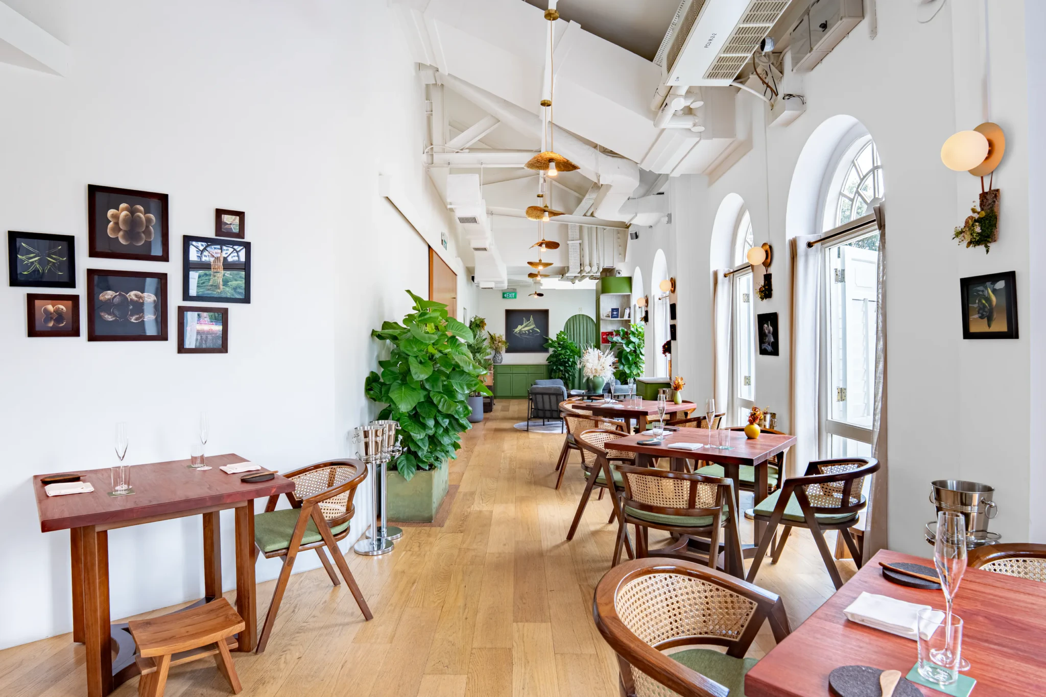 Restaurant JAG's dining room with wood tables, rattan chairs, framed artwork on white walls, arched windows, pendant lighting, and potted greenery