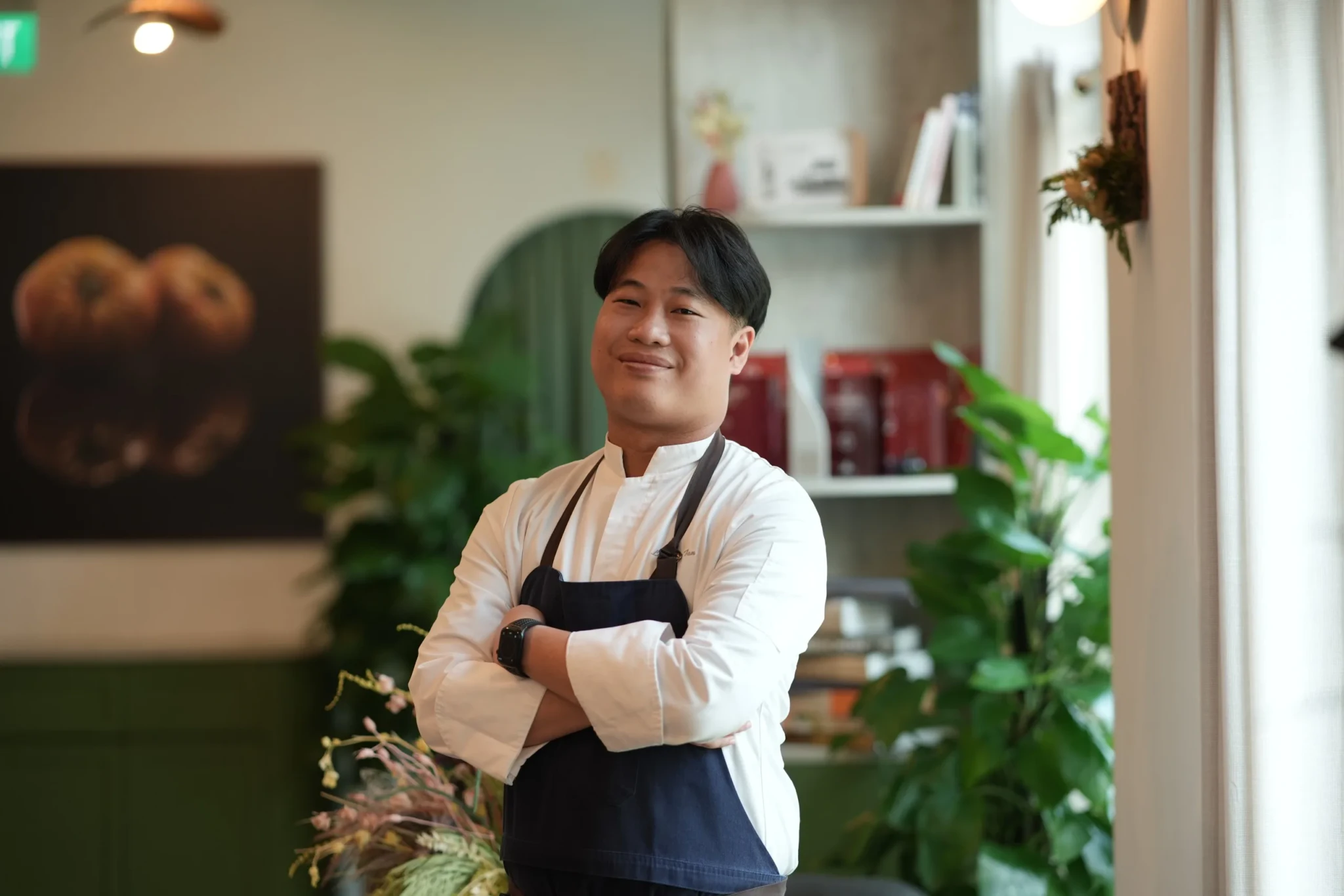 Chef Laurence Tan in white shirt and black apron, arms crossed, smiling in restaurant with plants and shelving