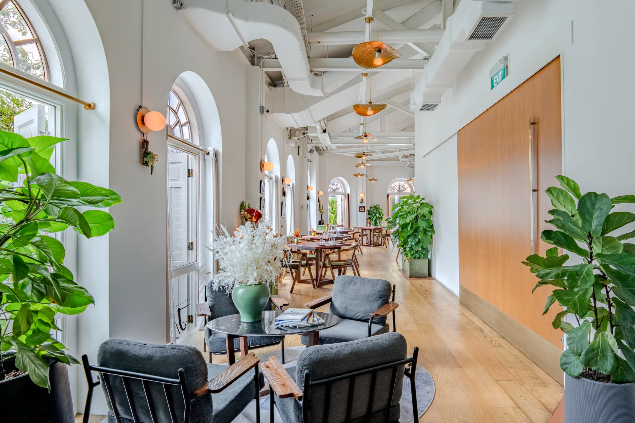 A bright corridor at Restaurant JAG with archway windows, green plants, amber pendant lights, and dining tables in the background