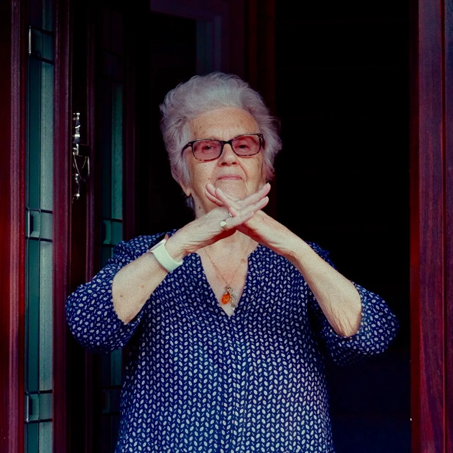 Vintage-style color photograph of an older woman wearing red-framed glasses and a patterned blue dress, arms crossed in an X gesture