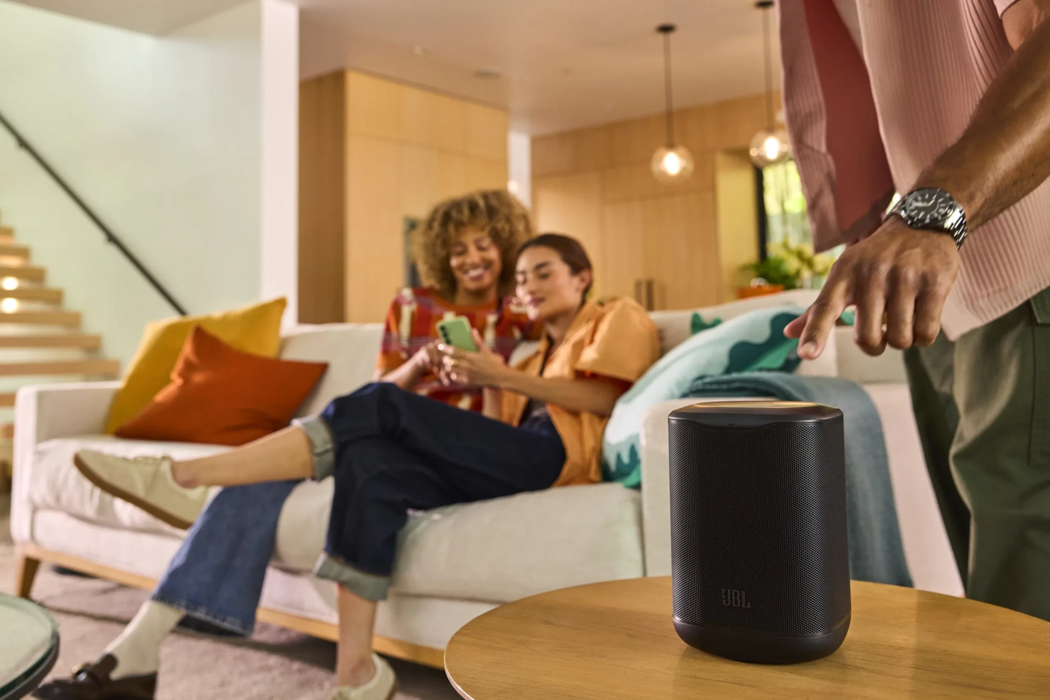 The JBL Cove M1 Wi-Fi speaker on a wooden table in a living room while three people sit on a nearby sofa