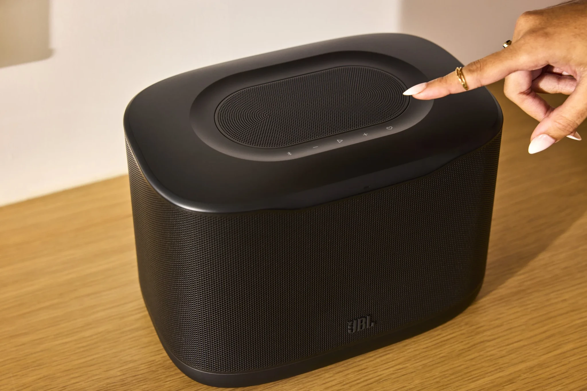 A hand reaching for the top of a black JBL Cove X1 speaker with mesh panels and touch controls on a wooden table