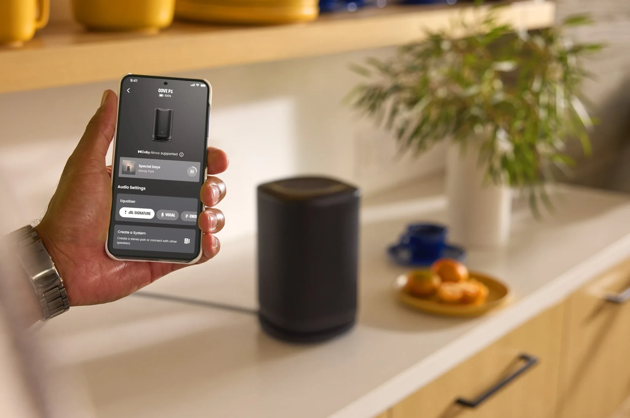 Hand holding smartphone displaying JBL Cove app with speaker visible on kitchen counter behind it
