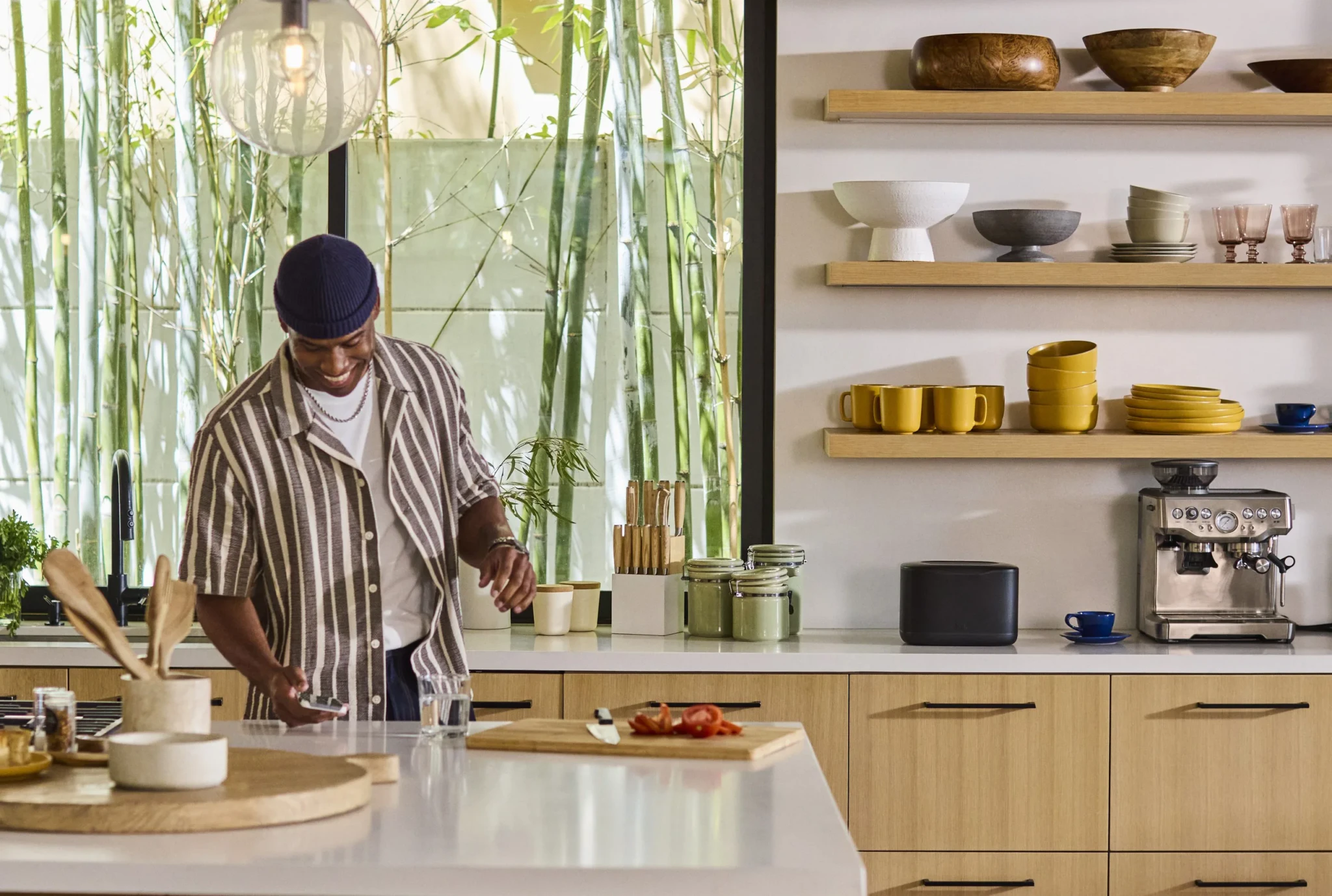 A man prepares food in a modern kitchen with JBL Cove speakers placed on open shelving beside dishware and a coffee machine