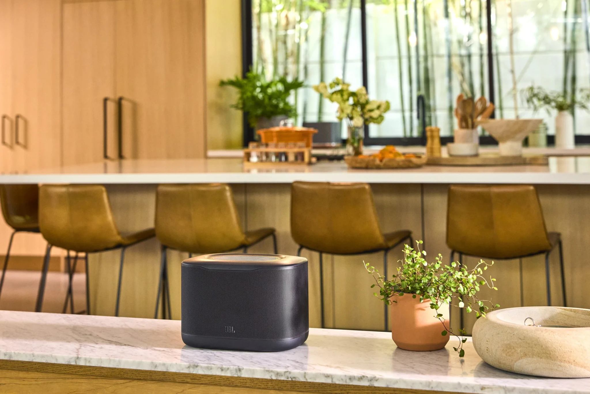 The JBL Cove X1 speaker on a marble kitchen counter with olive-green bar seating, potted plants and large windows