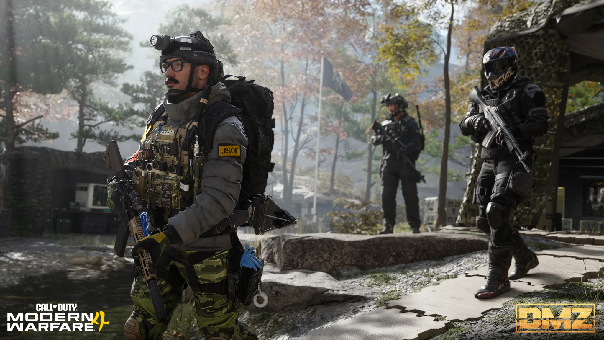 An operator in a grey jacket with a JSOF patch, glasses, and a headlamp-mounted helmet walking through the wooded Forward Operating Base with two armed operators behind him