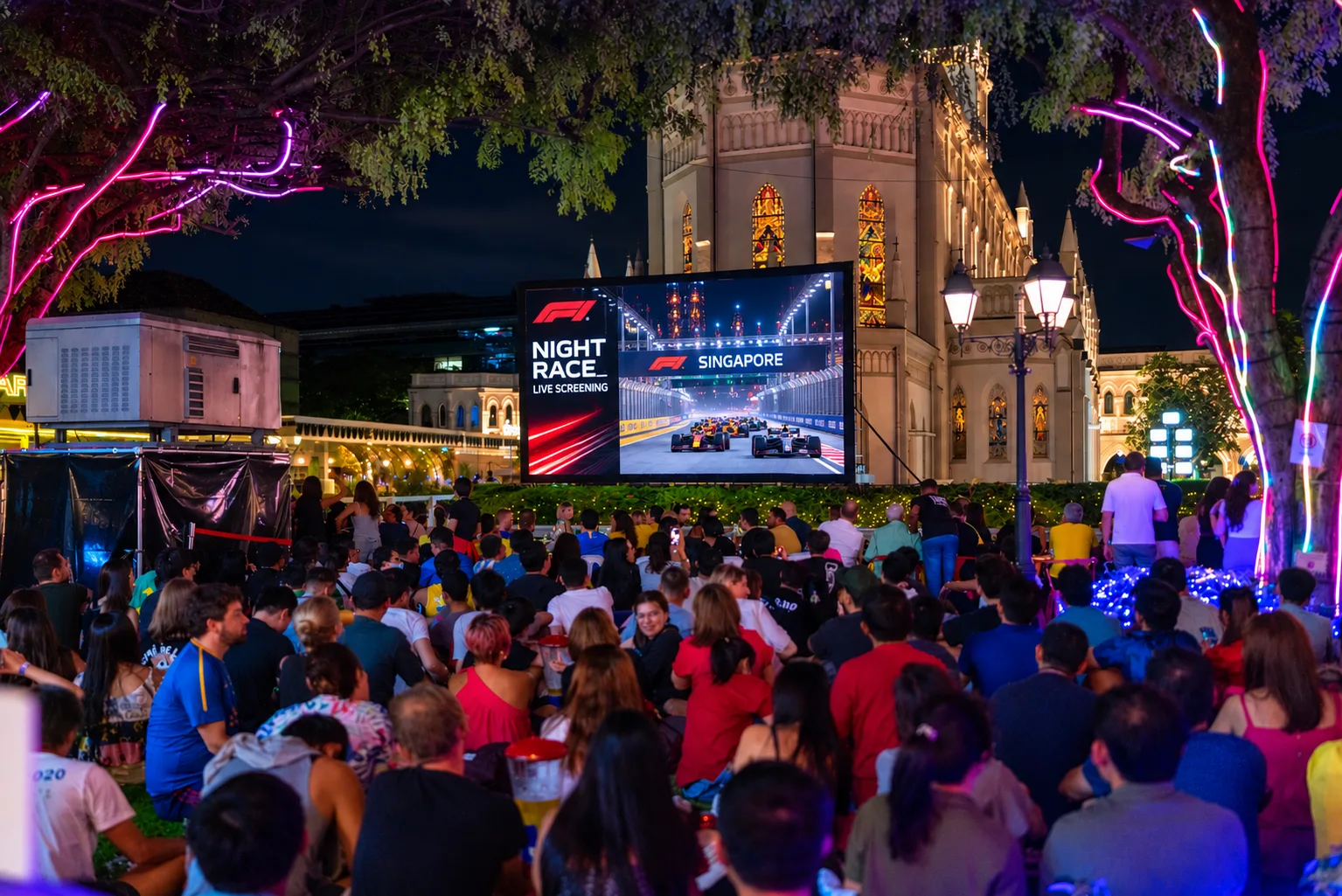 Crowd seated on grass watching a Singapore night race live screening on a large outdoor screen beside CHIJMES at night
