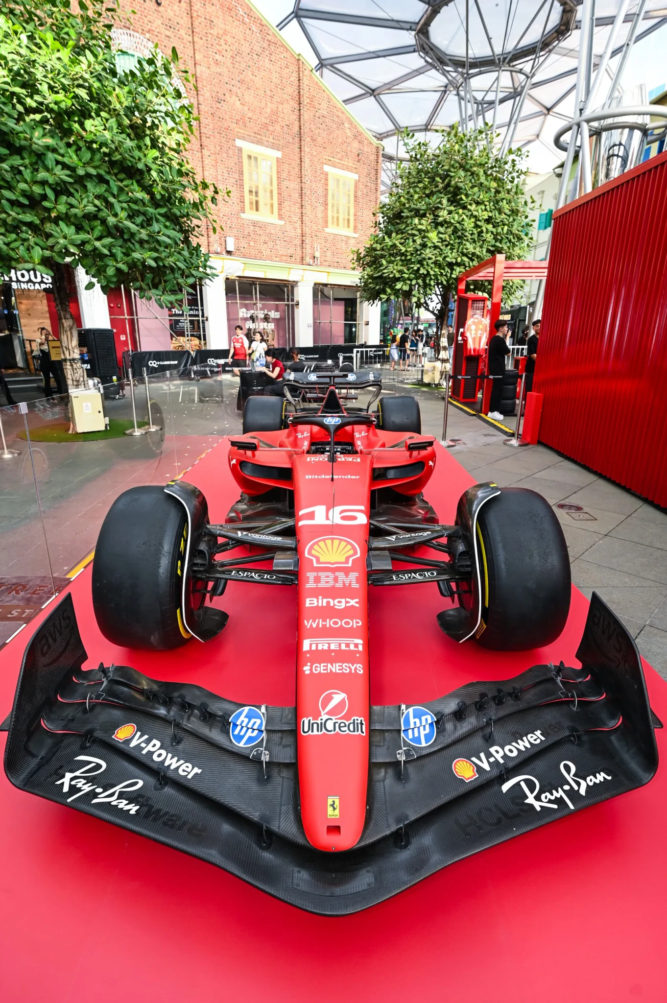 A Ferrari Formula 1 car numbered 16, carrying Ray-Ban and other sponsor branding, displayed in a public courtyard