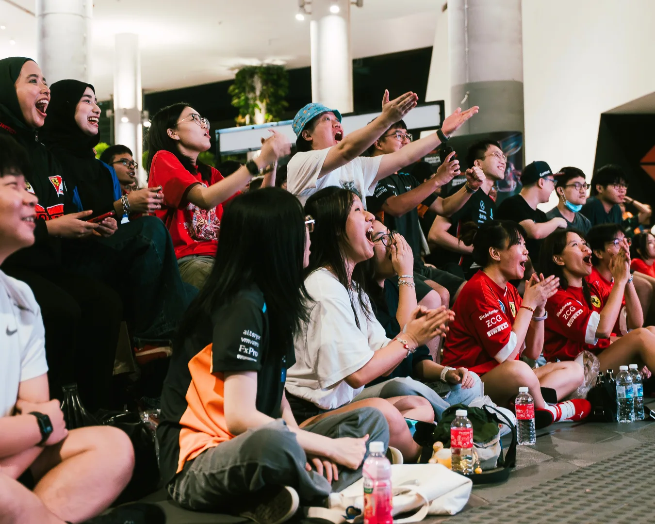 Fans seated on the floor of a mall atrium cheering and clapping at a live F1 screening, many in Ferrari and Mercedes shirts