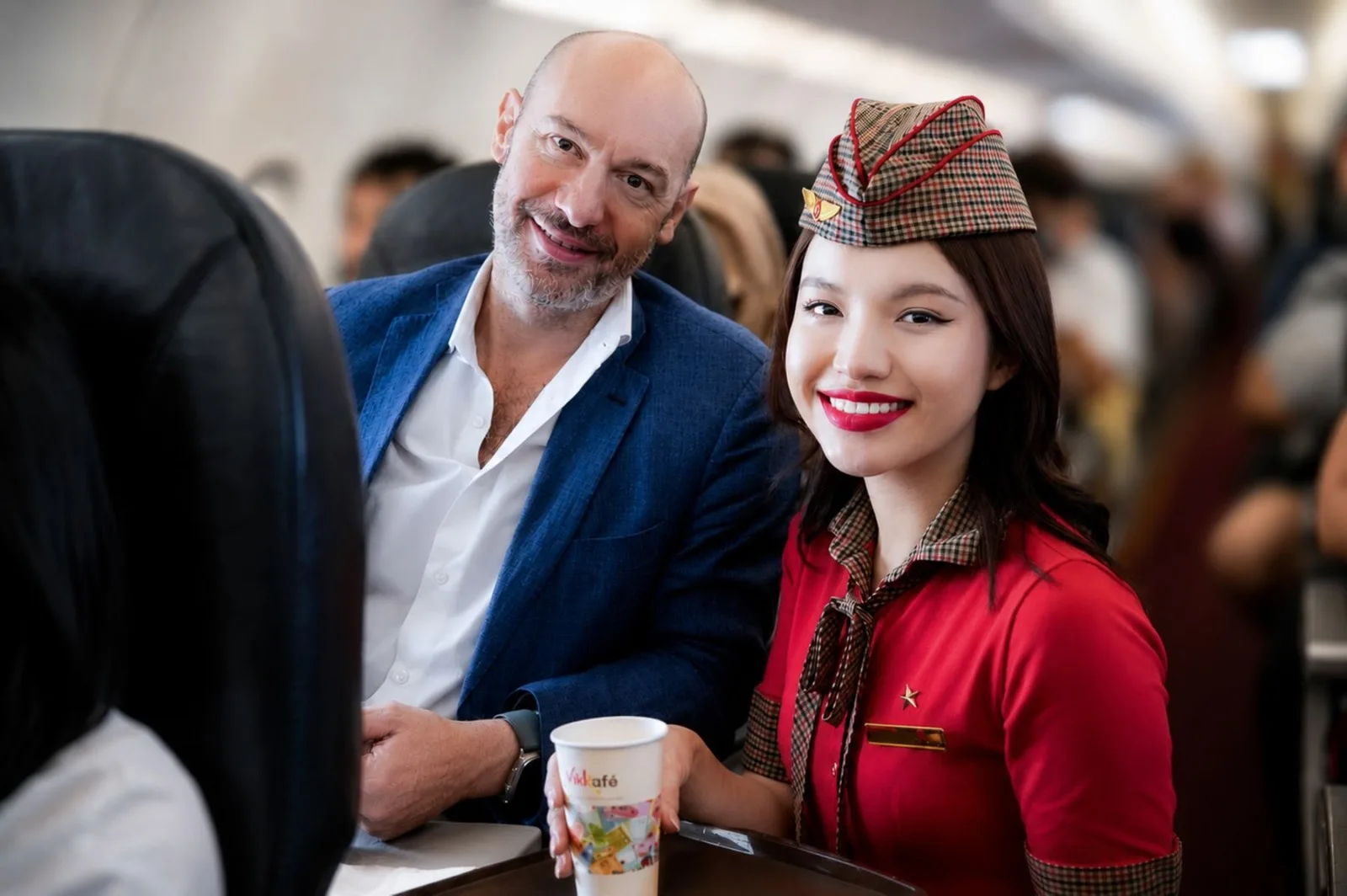 A Vietjet cabin crew member in a red uniform serving a seated passenger aboard an aircraft