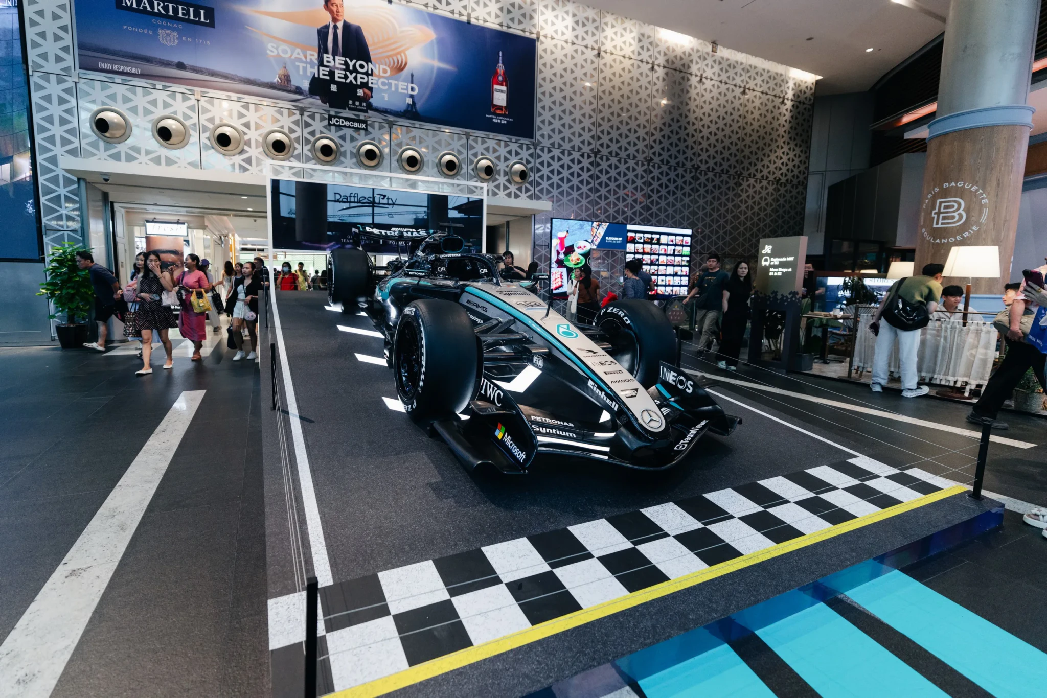 Black and teal Mercedes F1 car displayed indoors at a mall pop-up with checkered floor and crowds