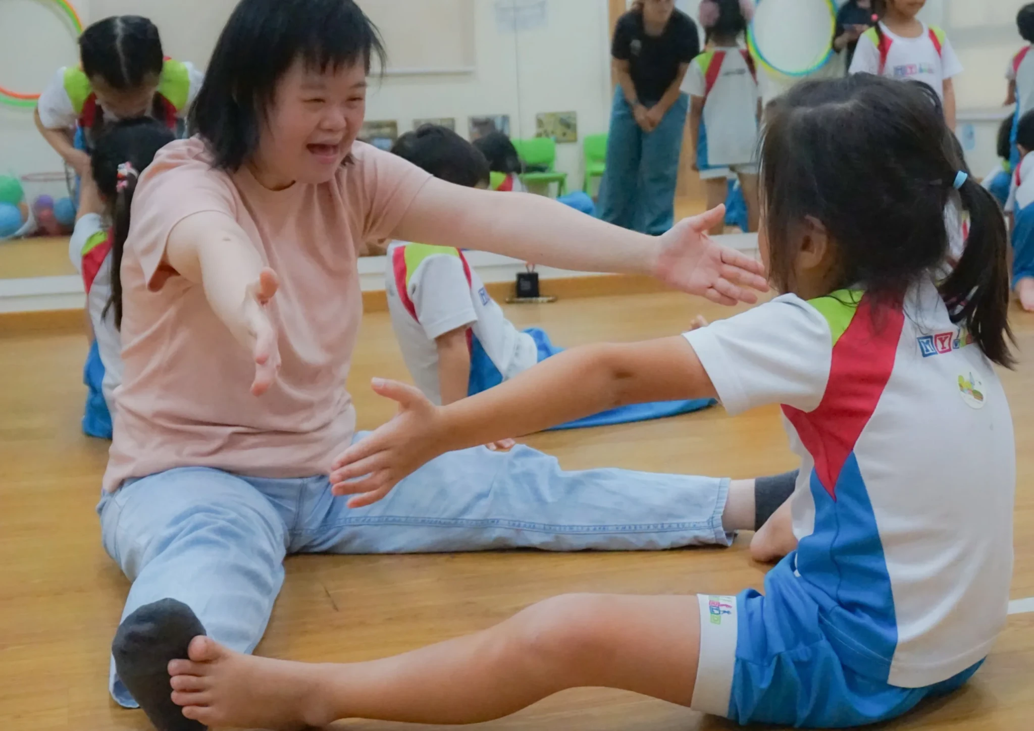 An Assistant Movement Co-facilitator guides a young preschooler through a stretching exercise on a classroom floor