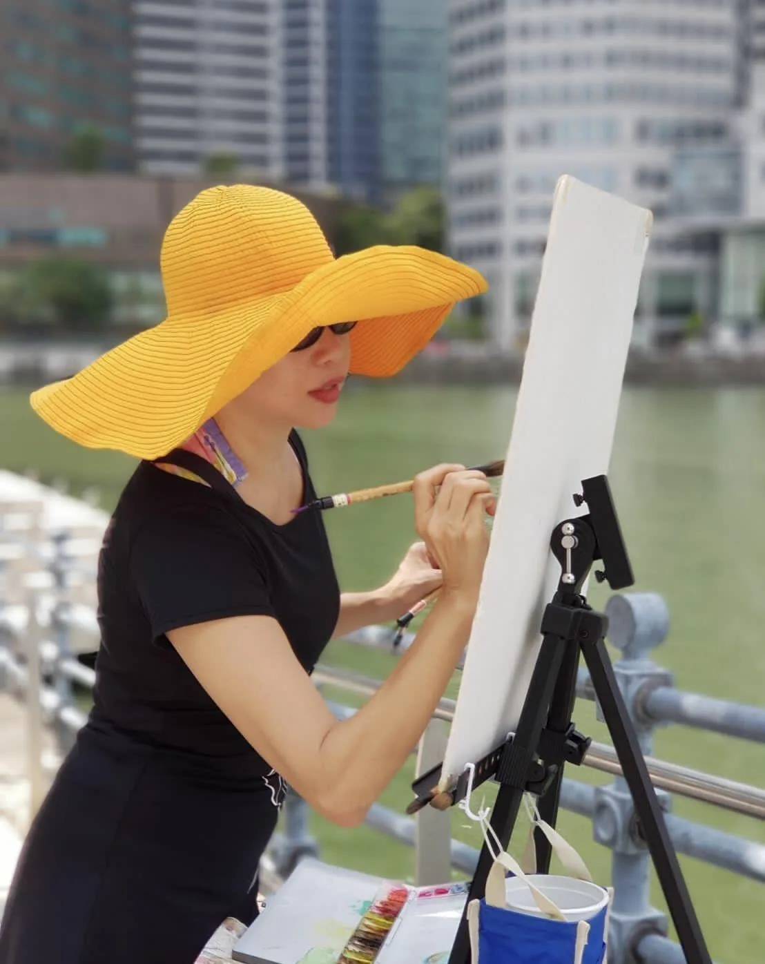 A watercolour artist in a wide-brimmed yellow hat paints at an easel beside a river, with city buildings behind her