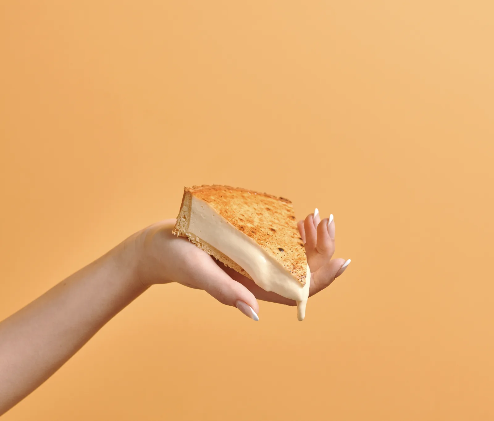 A slice of Olivia's Signature Cheesecake with a golden-brown crust, held in a hand against a warm yellow background