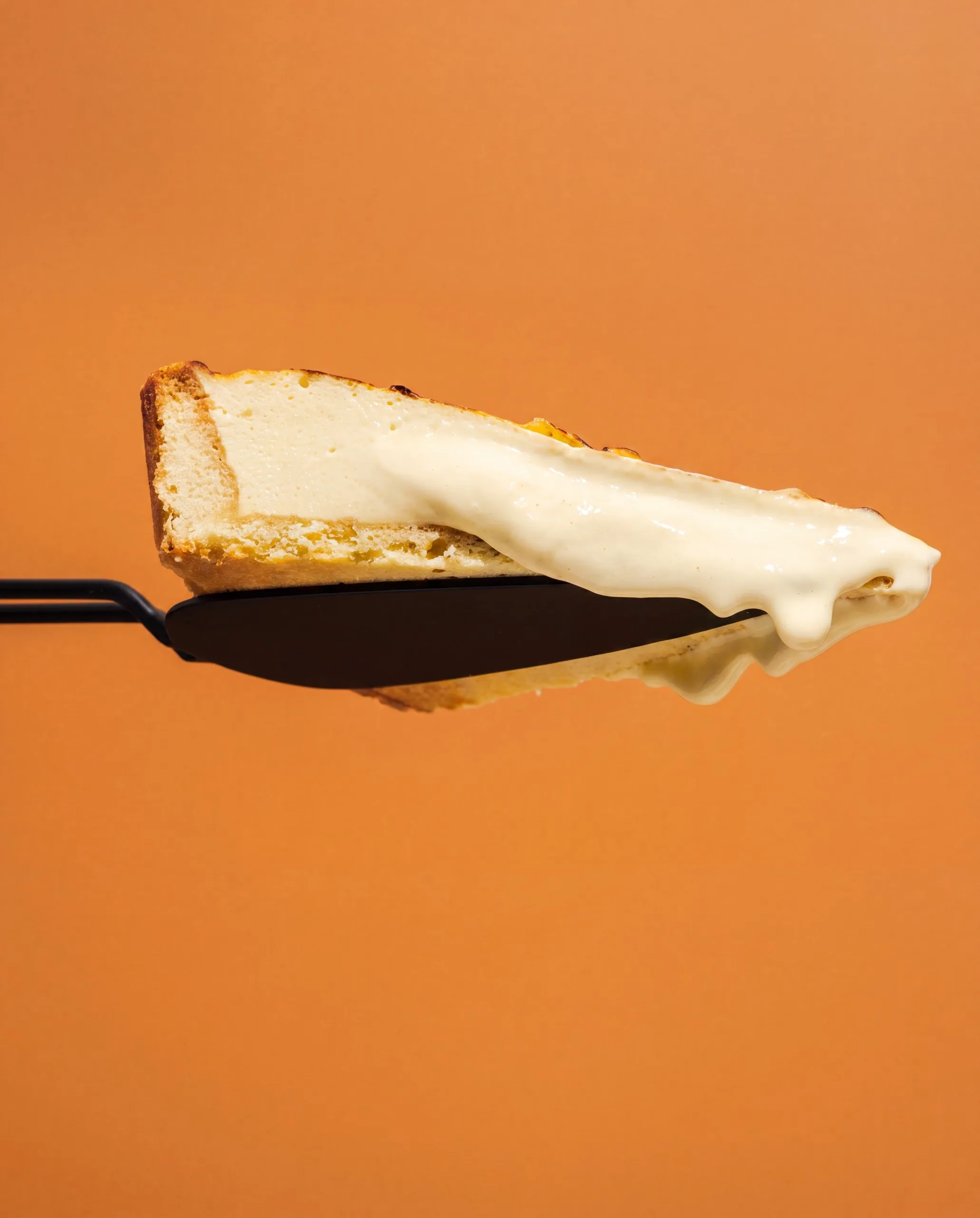A creamy wedge of Olivia's Signature Cheesecake on a dark spreader against an orange background