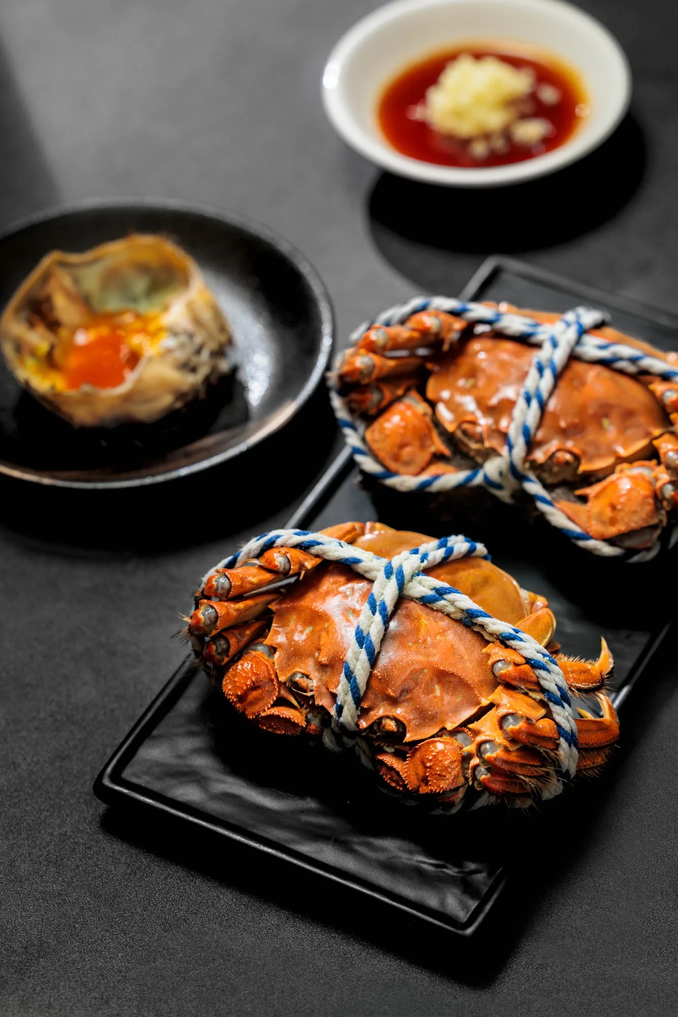 Hairy crabs tied with blue and white twine on a black rectangular plate, with a pan-seared dish and dipping sauce in the foreground