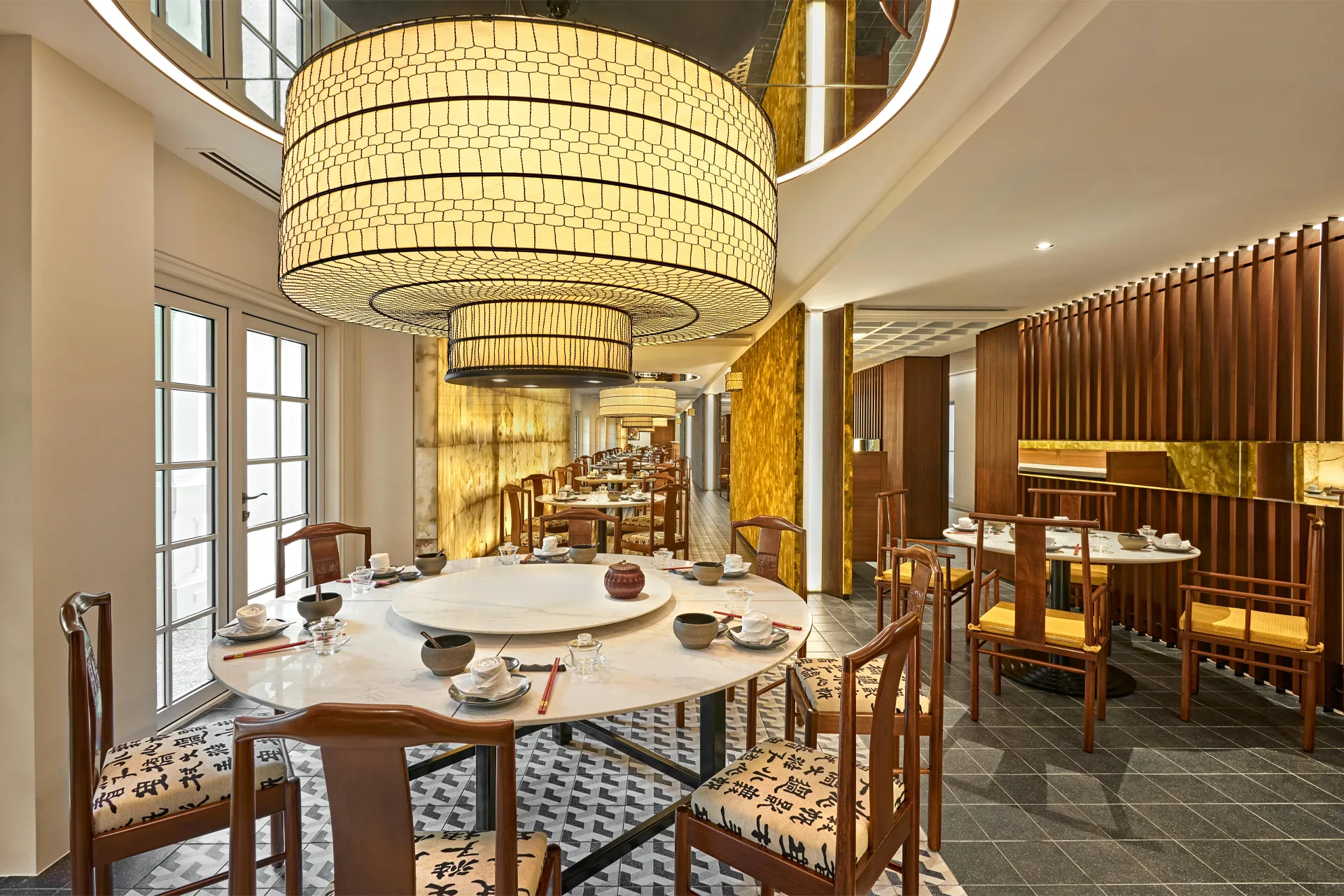 Main dining hall at Min Jiang with a large cylindrical woven pendant lamp, round marble-topped tables, and wooden chairs