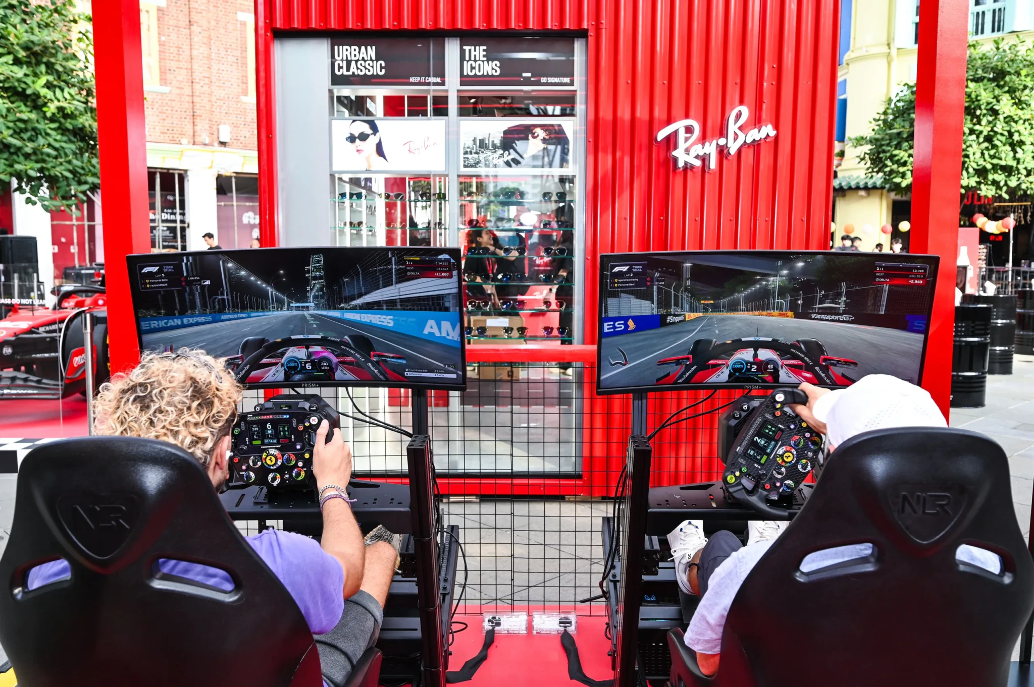 Two people seated at racing simulators with a Ray-Ban storefront behind them
