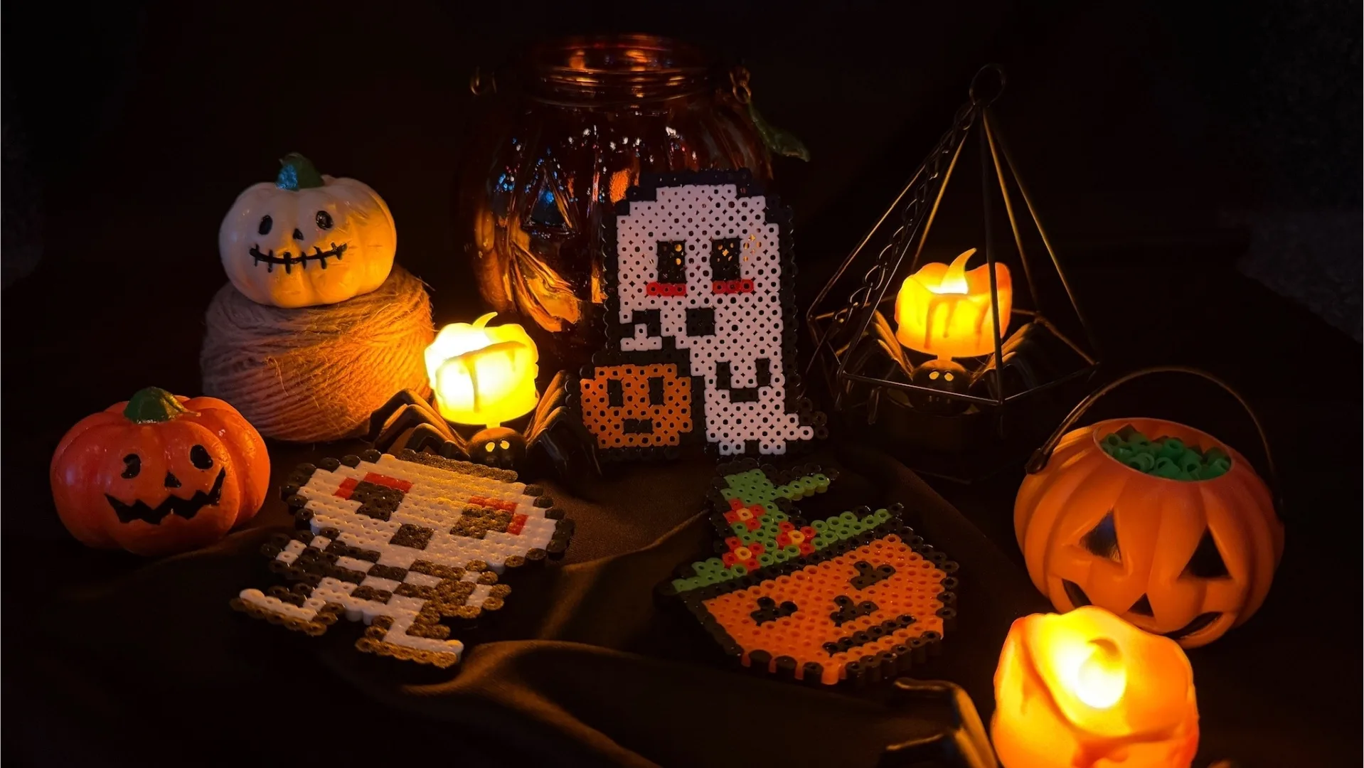 Hama bead artworks of jack-o'-lanterns, ghosts, and pixel-art Halloween characters displayed beside carved pumpkins and lit lanterns