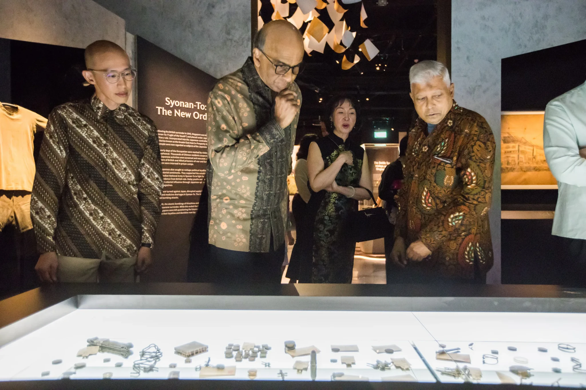 Visitors look over artefacts in a lit display case beneath a Syonan-To panel in a museum gallery.