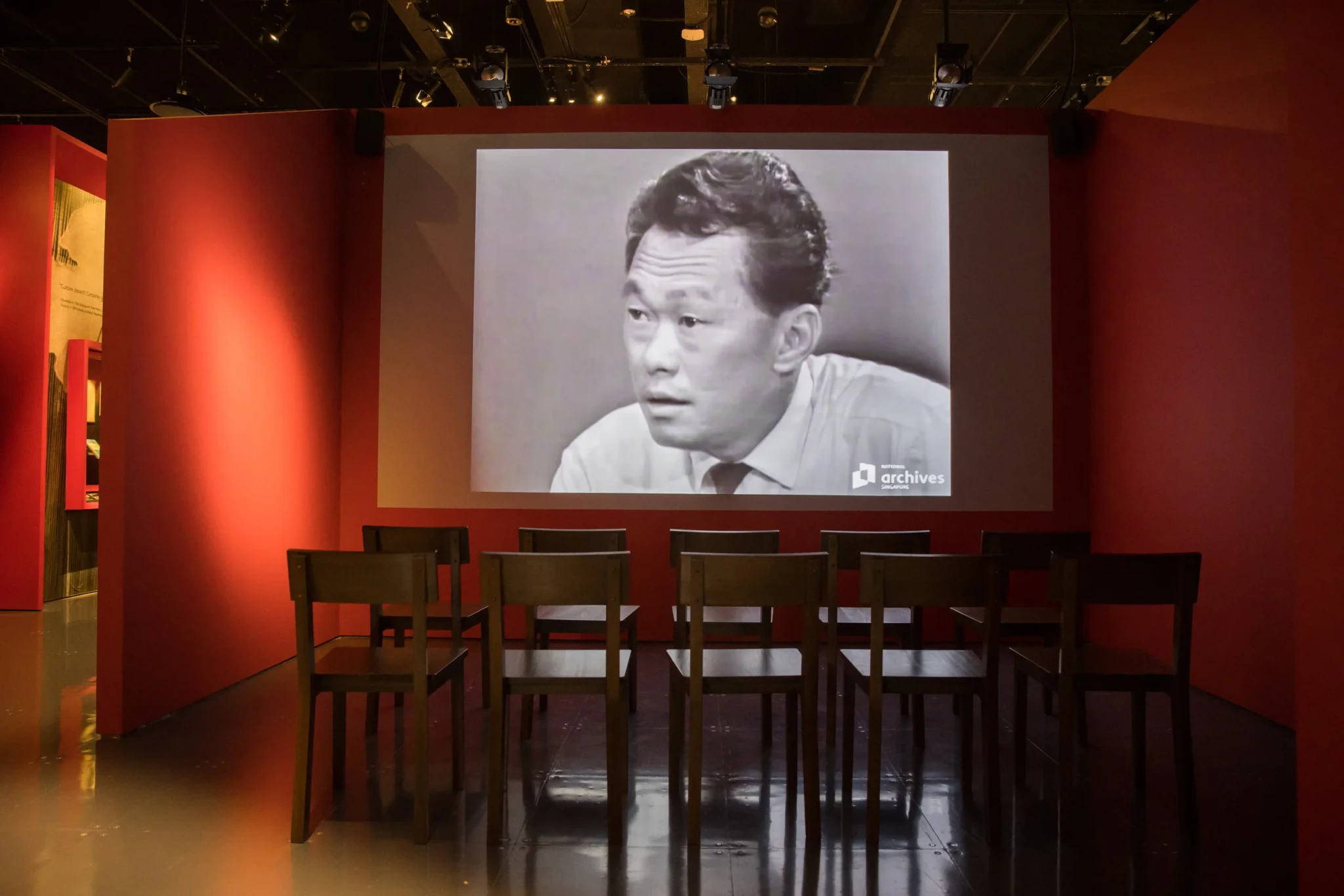 Archival film footage projected on a red wall above rows of wooden chairs in the Divergent Paths display