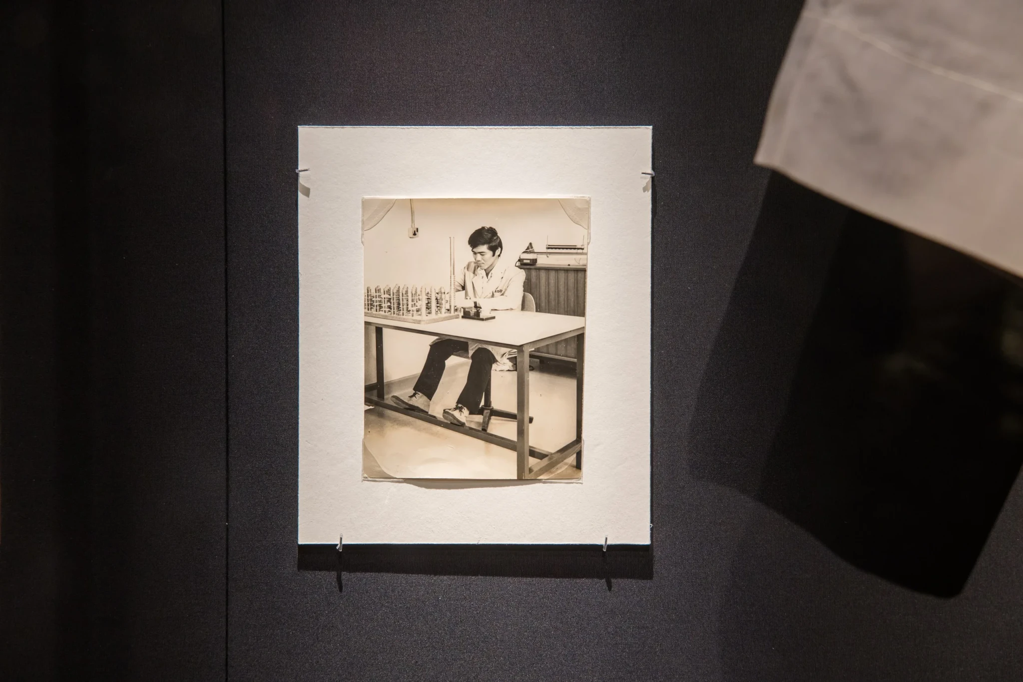 A sepia photograph of Mr Chong Nam Soy seated at a table assembling camera shutters, framed on a dark wall