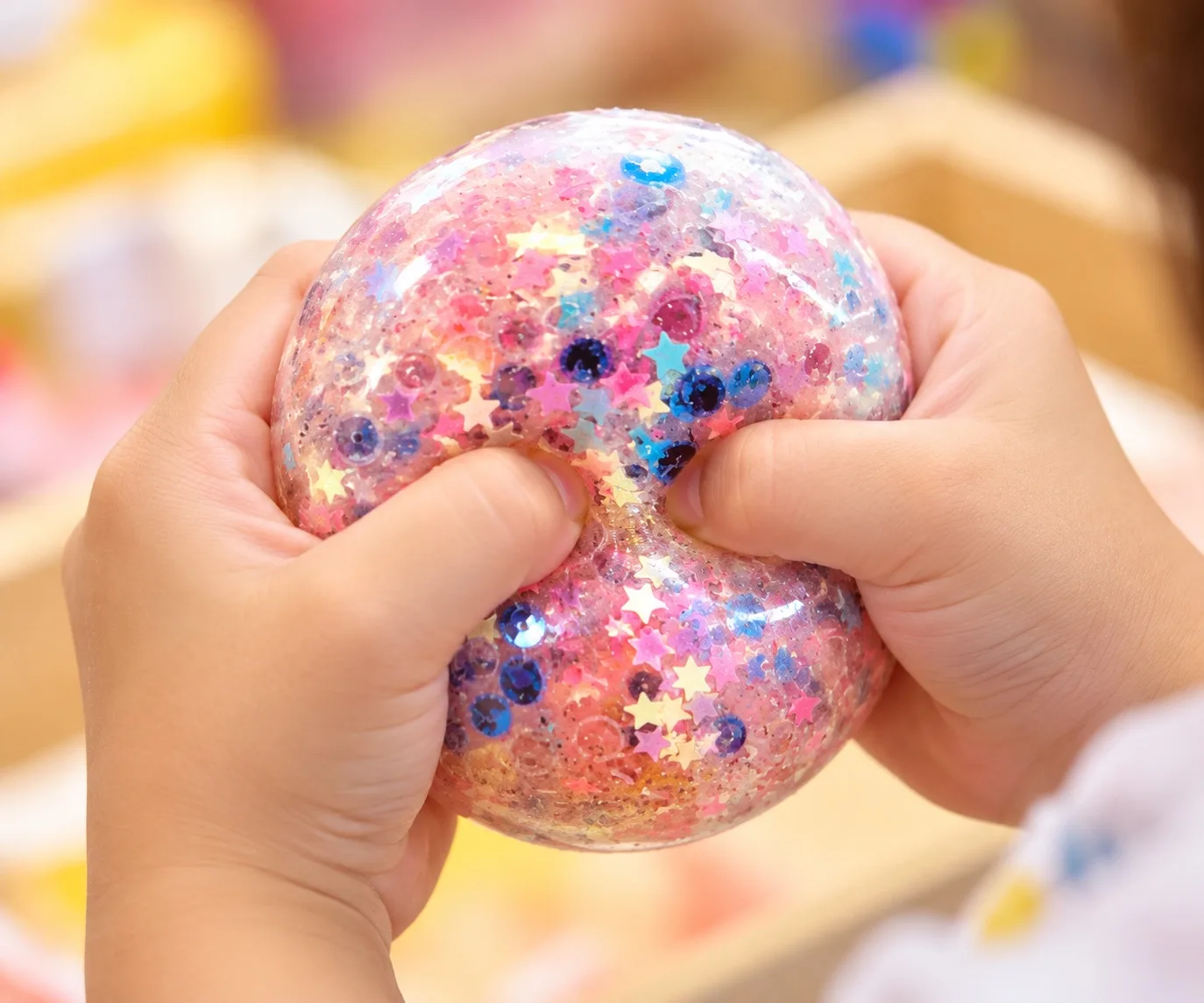 Two hands squeezing a clear glitter stress ball filled with pink, blue and star-shaped sequins