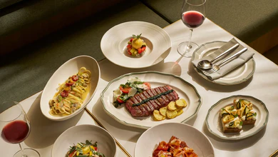 A table spread of Osteria Da Su dishes with clam spaghetti, farfalle in tomato sauce, bruschetta, salad, sliced steak and red wine
