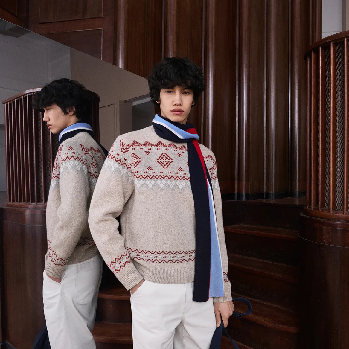 Models in a cream Fair Isle knit sweater and a navy and light blue striped scarf in front of a wood-panelled staircase