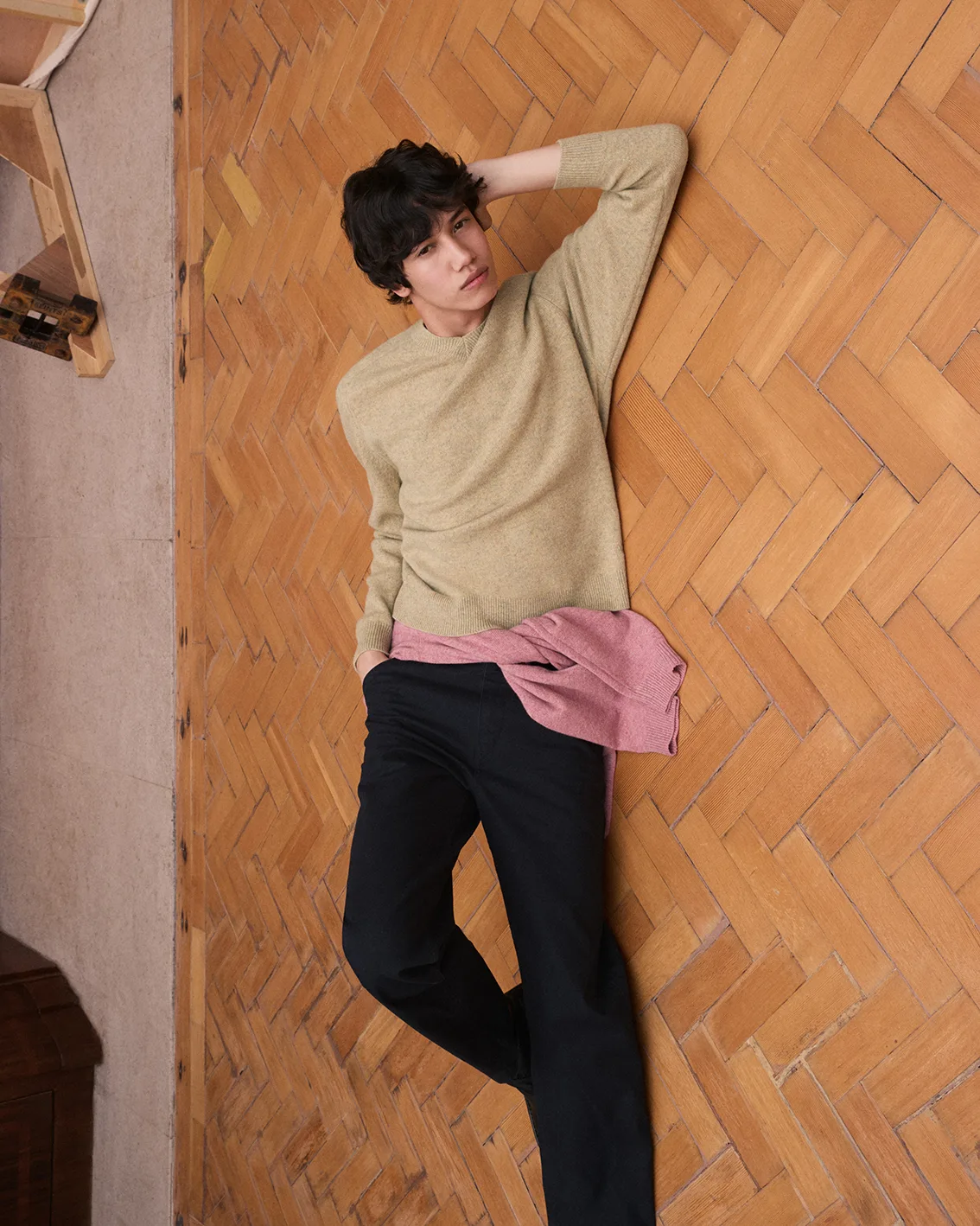 Model in a sage knit crewneck with a pink knit tied at the waist and dark navy trousers, posed against a herringbone parquet wall