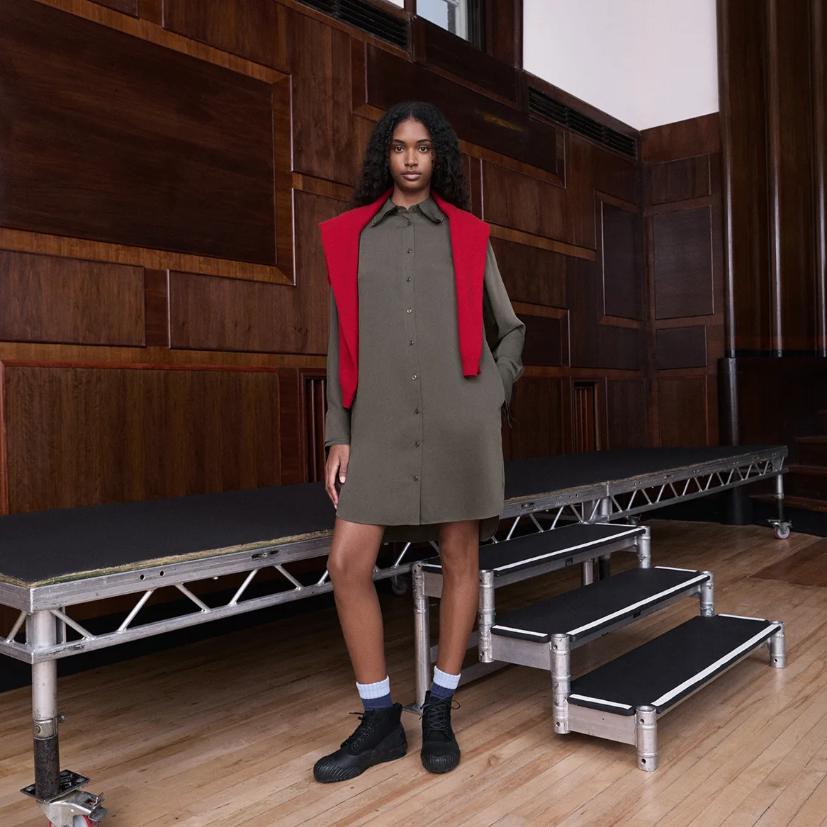 Model in the UNIQLO and JW ANDERSON olive shirt dress with a red knit draped over her shoulders, beside silver staging in a wood-panelled room