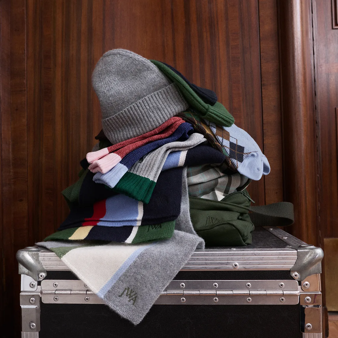 UNIQLO and JW ANDERSON knitwear and accessories stacked on a metal flight case, including a grey beanie, striped scarves, an argyle sweater and a green bag