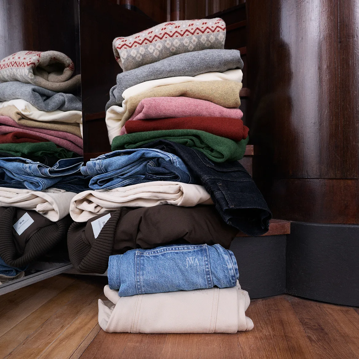 Folded UNIQLO and JW ANDERSON knitwear and denim stacked on a wooden floor in front of a dark wooden cabinet