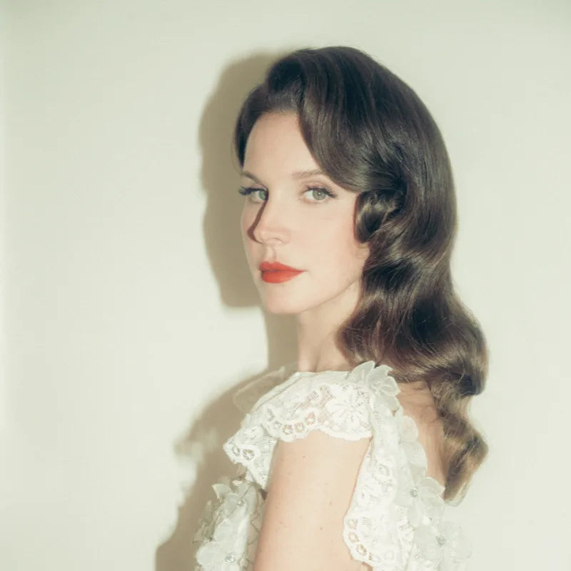 Lana Del Rey in profile with dark wavy hair, red lipstick and a white lace dress, looking at the camera over her shoulder