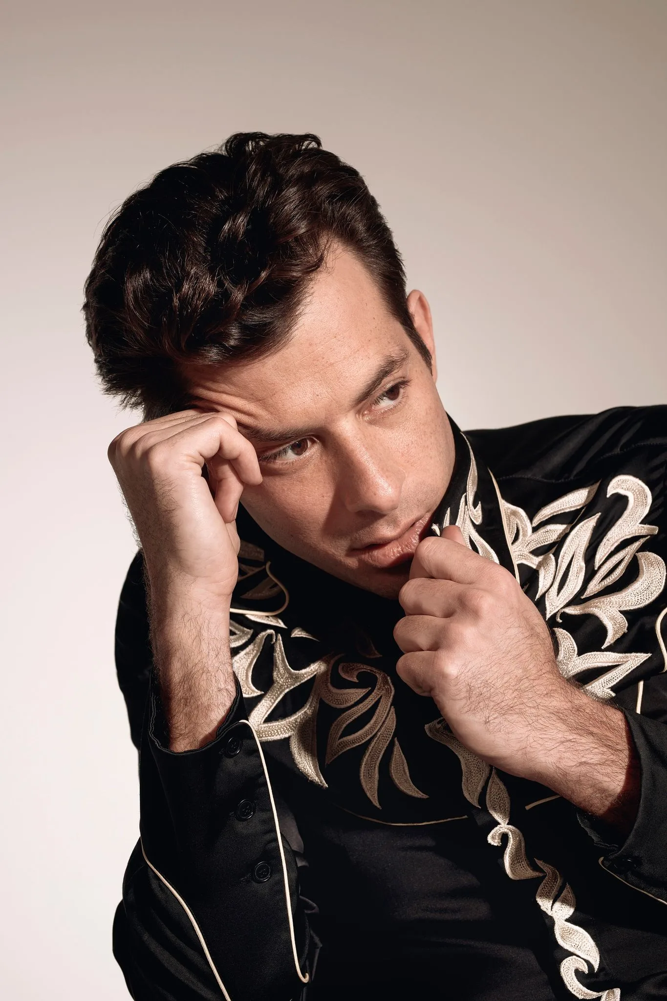 Mark Ronson with dark swept-back hair and a black embroidered western shirt, one hand to his temple, looking aside