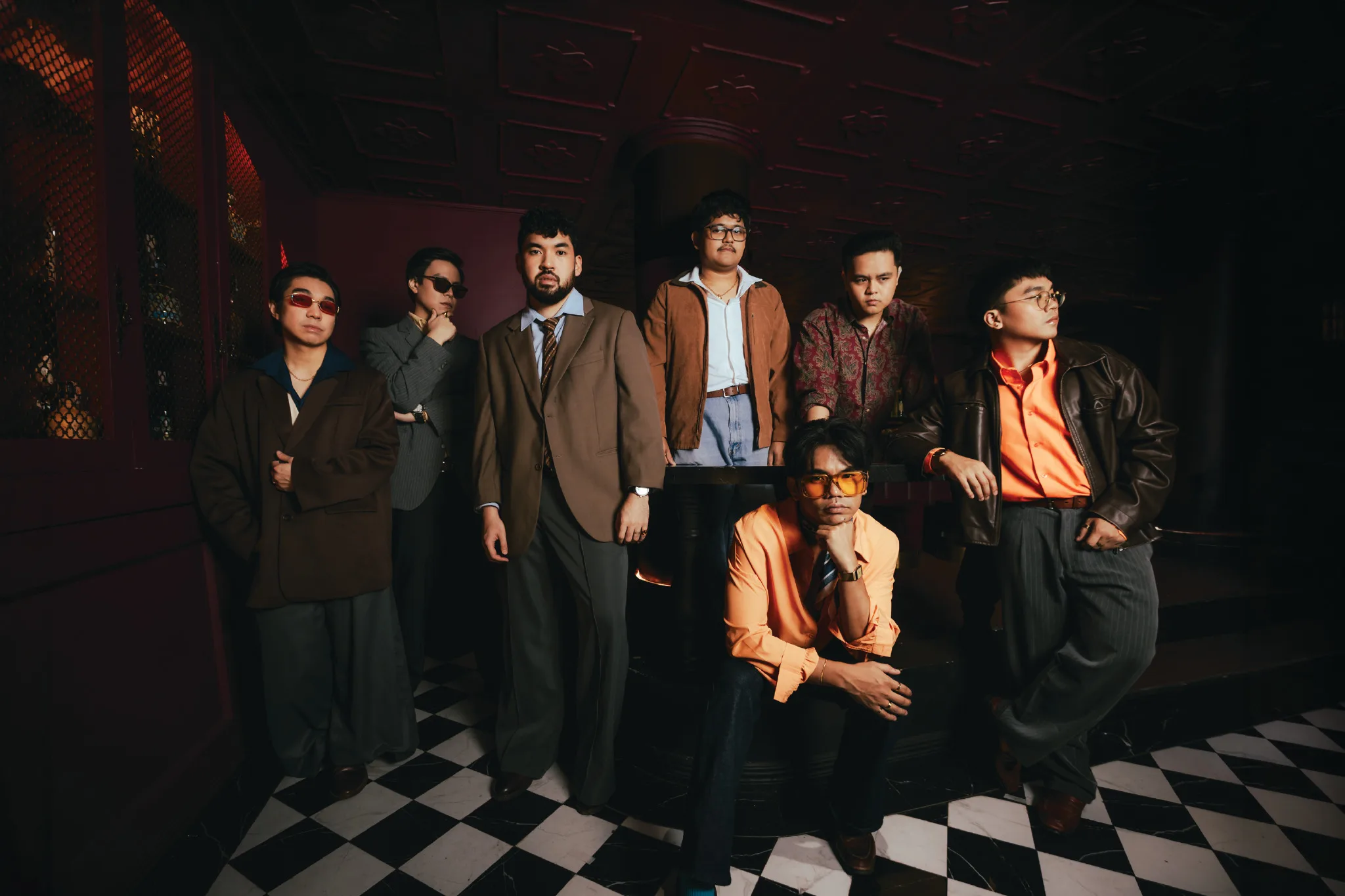 The seven members of Lola Amour in vintage suits and jackets posing in a dark red room with a black and white checkered floor