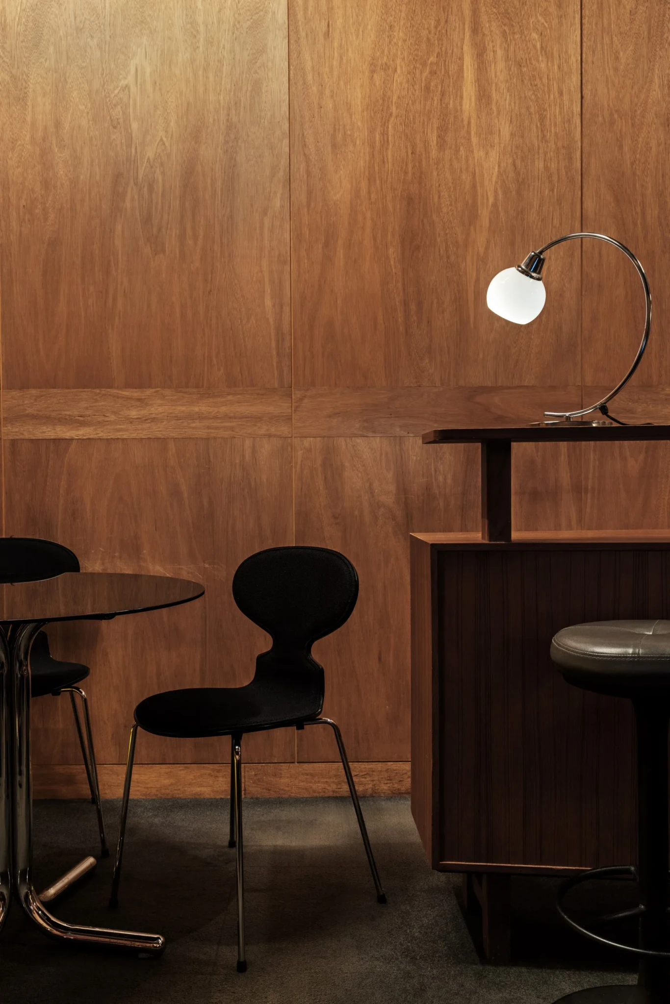 Warm wood-panelled room with black chairs at a round chrome-pedestal table, a bar counter, and a white globe lamp on a chrome arm.