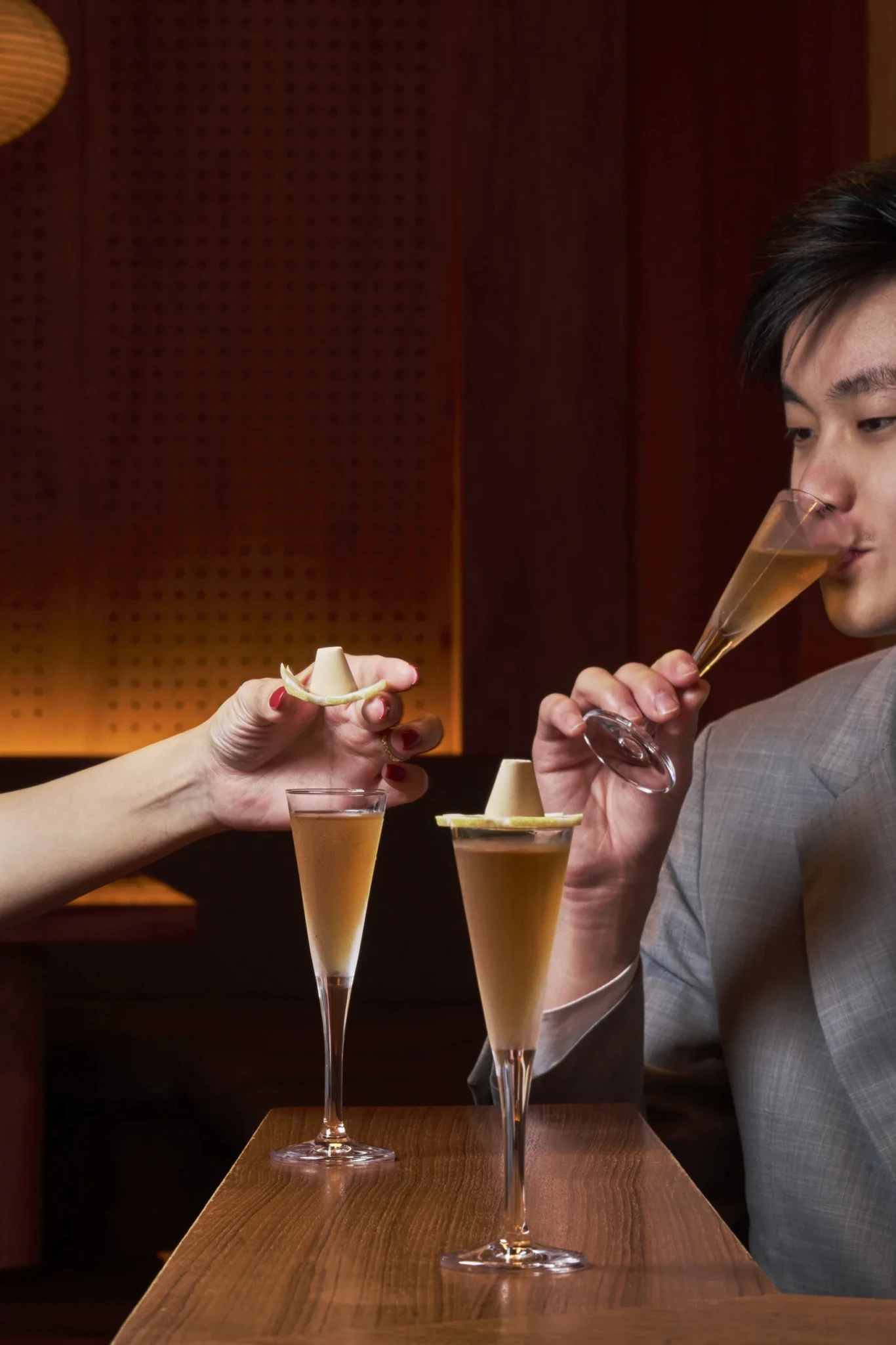 Man in grey suit sipping from a champagne flute as a woman's hand holds out a garnished cocktail glass