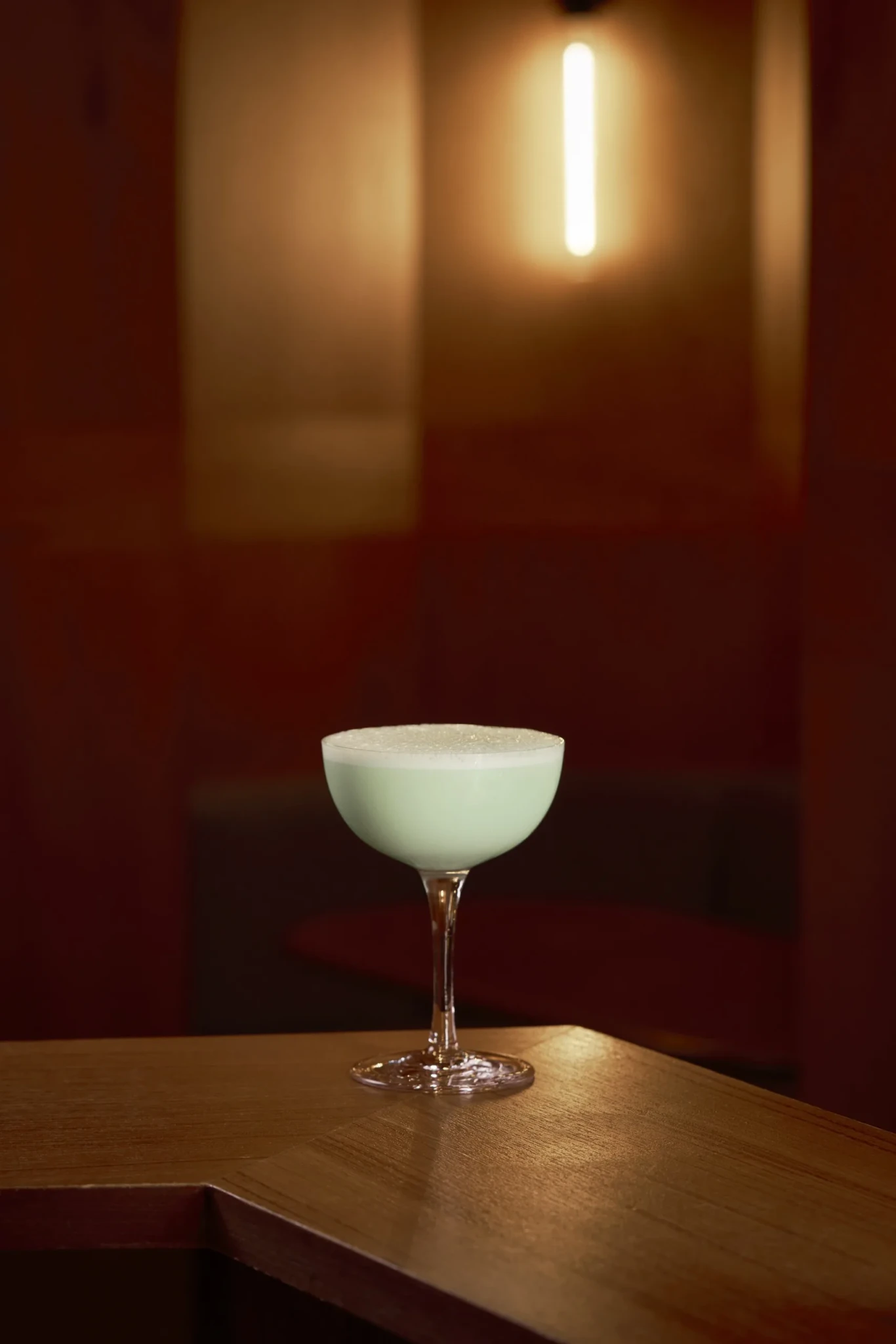 A pale green Grasshopper cocktail with a frothy top in a coupe glass on a wooden bar counter