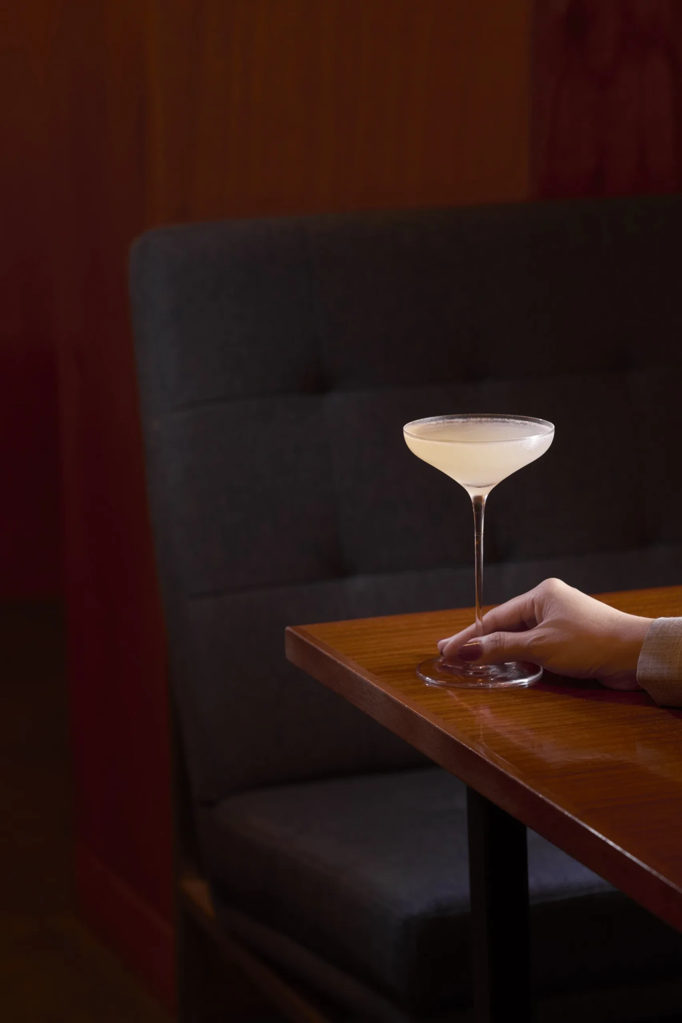 A Pear in Bloom cocktail with a frothy top in a coupe glass on a wooden table beside a dark banquette