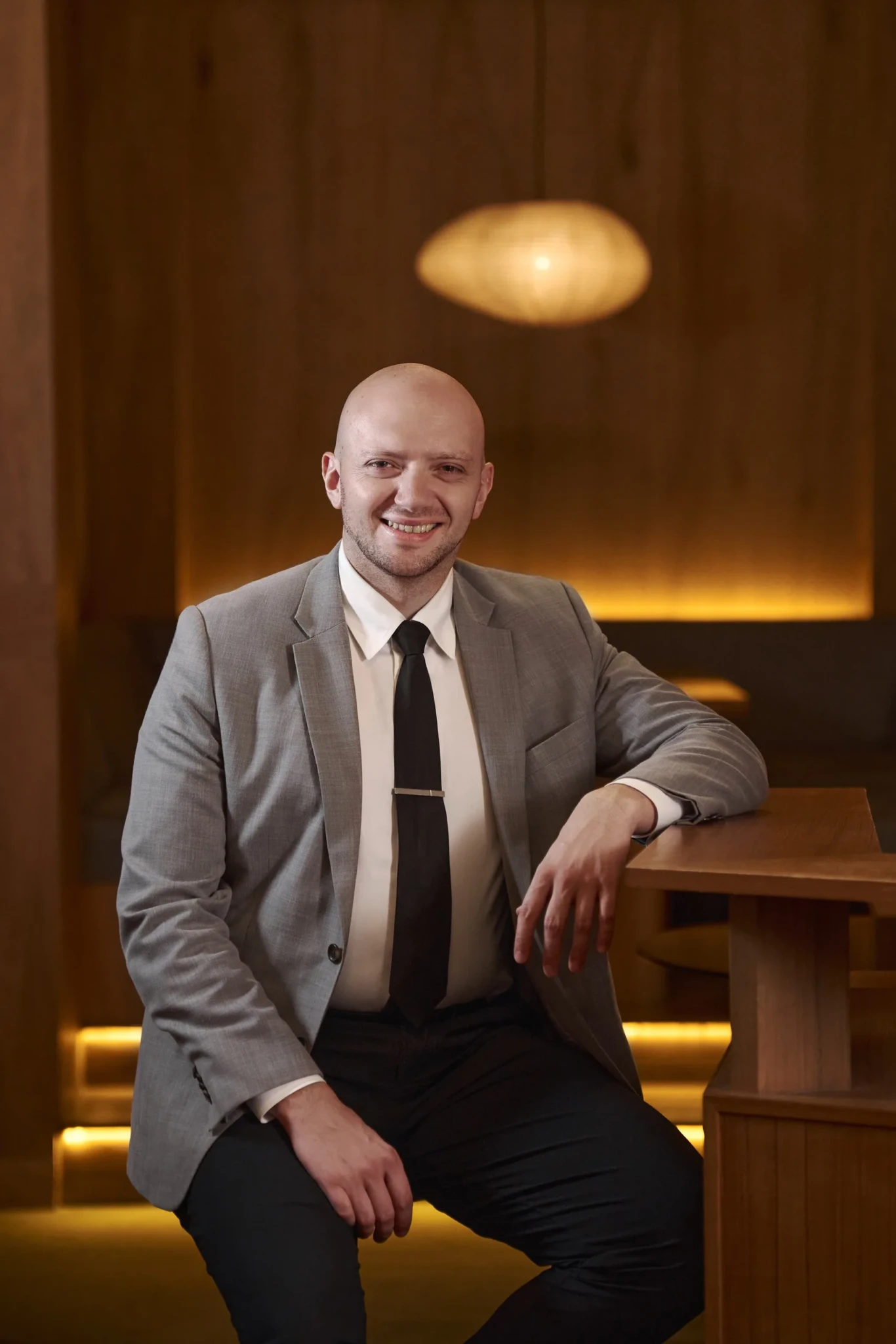 Luca Lulli, Principal Bartender at Live Twice, smiling at a wooden bar in a grey blazer and black tie