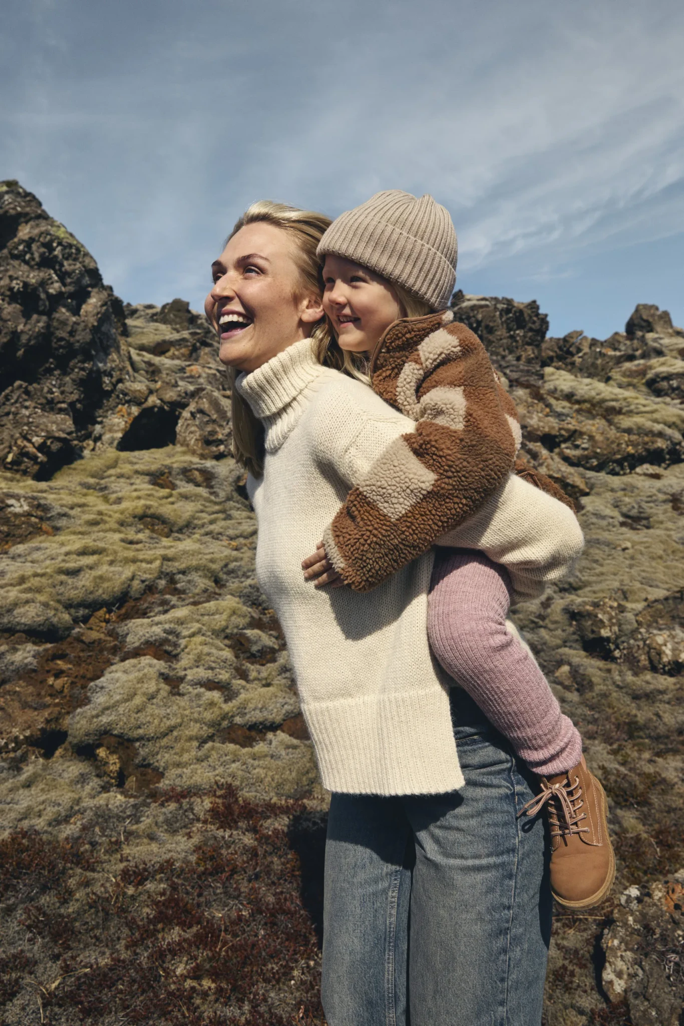 A woman carries a young child on her back across mossy volcanic rock under a blue sky