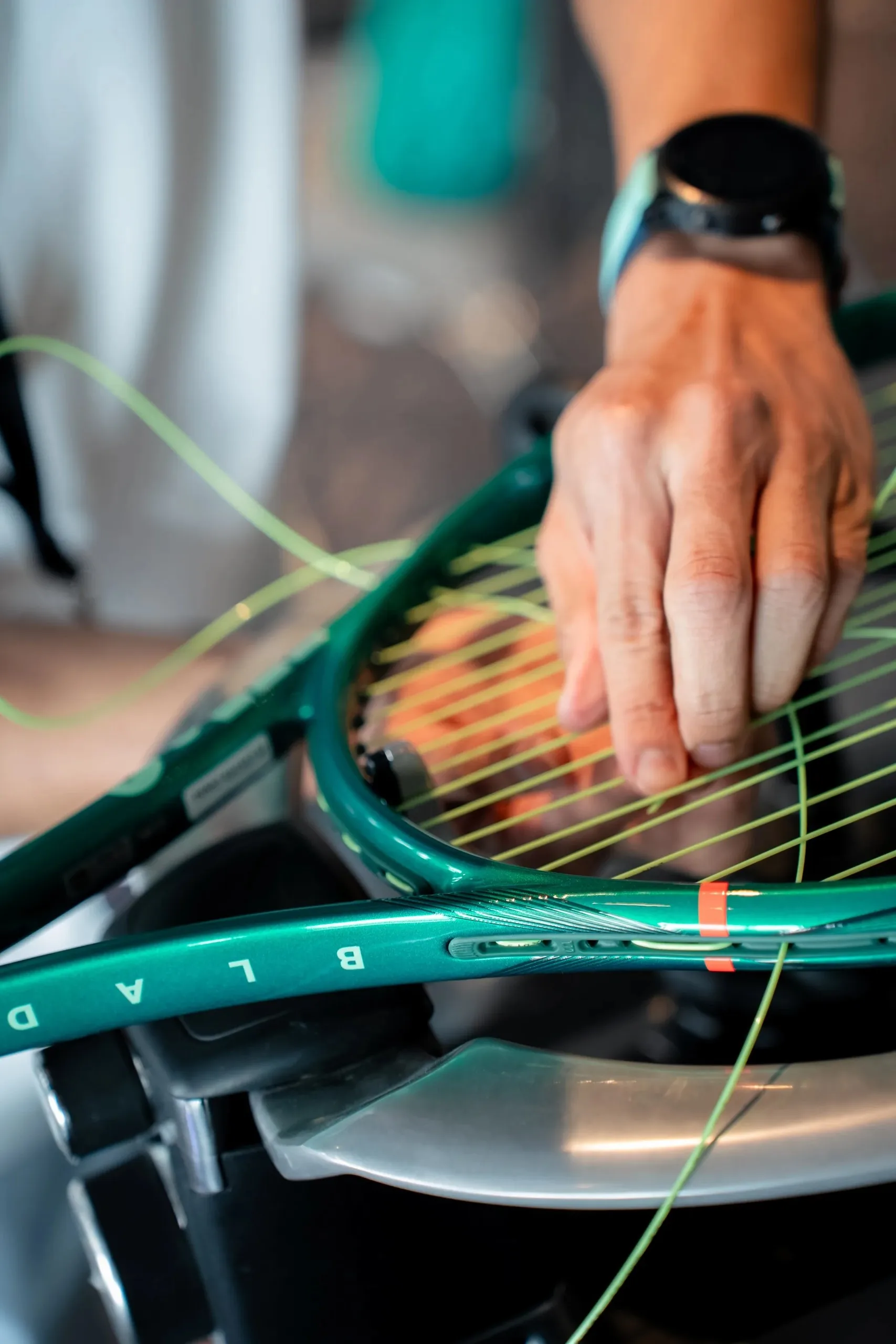 A hand threads green strings through a teal Wilson racket mounted on a stringing machine