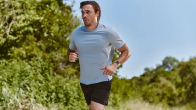 Man in a light blue t-shirt and black shorts running on a grassy trail past green trees, with a fitness tracker on his wrist.