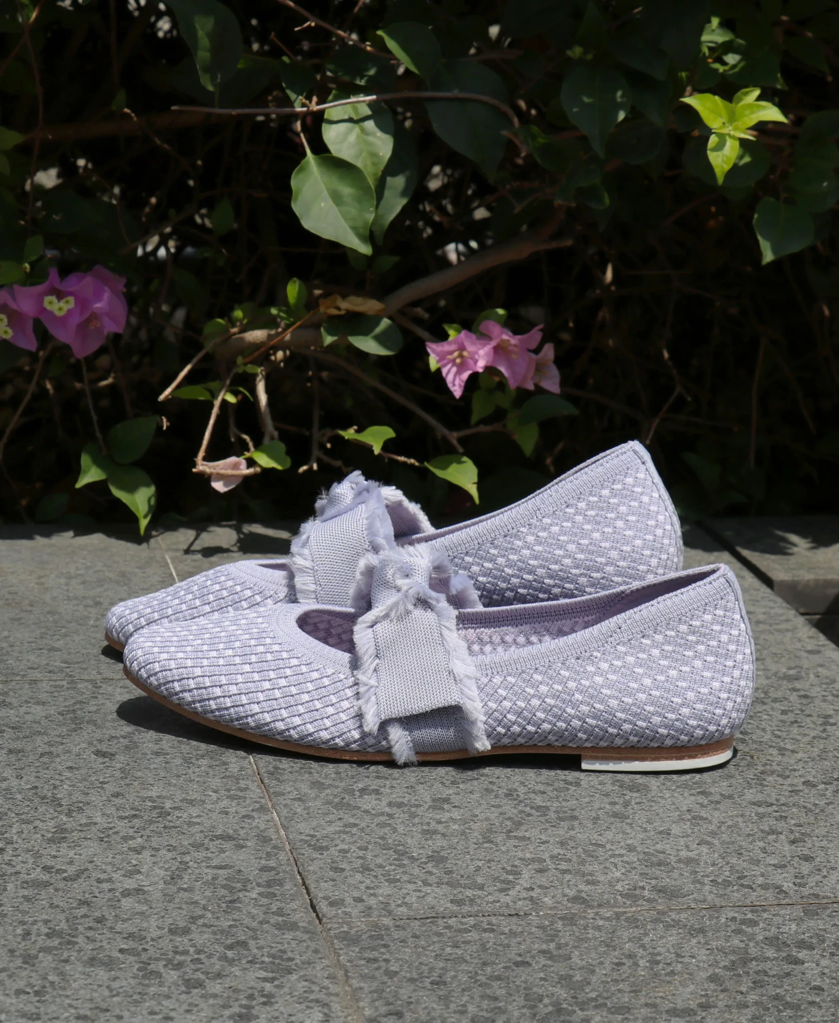 Lavender Skechers Cleo Snip flats with fringed ribbon detailing on stone paving beside pink bougainvillea