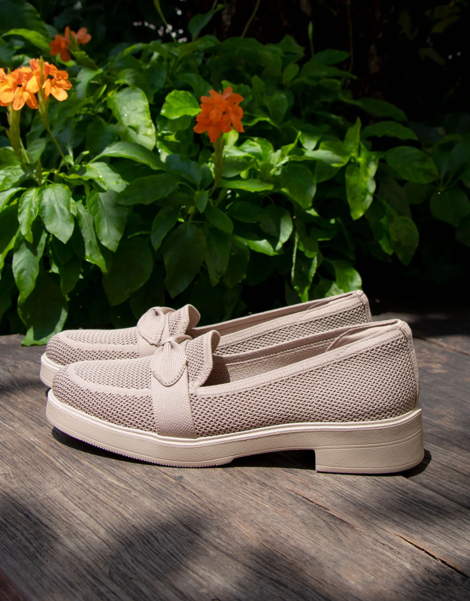 Taupe Skechers Cleo Taylor slip-on shoes with knit uppers and bow trim on a wooden table beside orange flowers