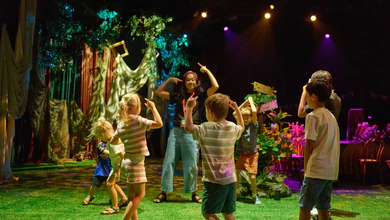 Children copying a facilitator's arm-pointing poses during a drama game on a forest-themed stage set