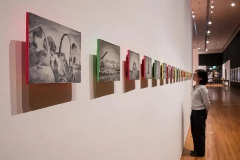 A visitor looks at a row of small framed grayscale drawings with coloured edges on a gallery wall