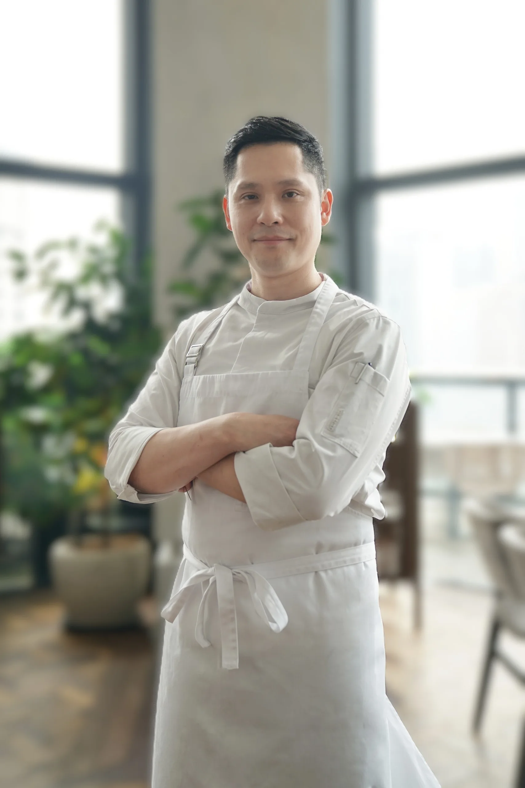 Chef Allen Suh of Restaurant Allen in a white chef's jacket and apron, standing in front of potted plants and tall windows